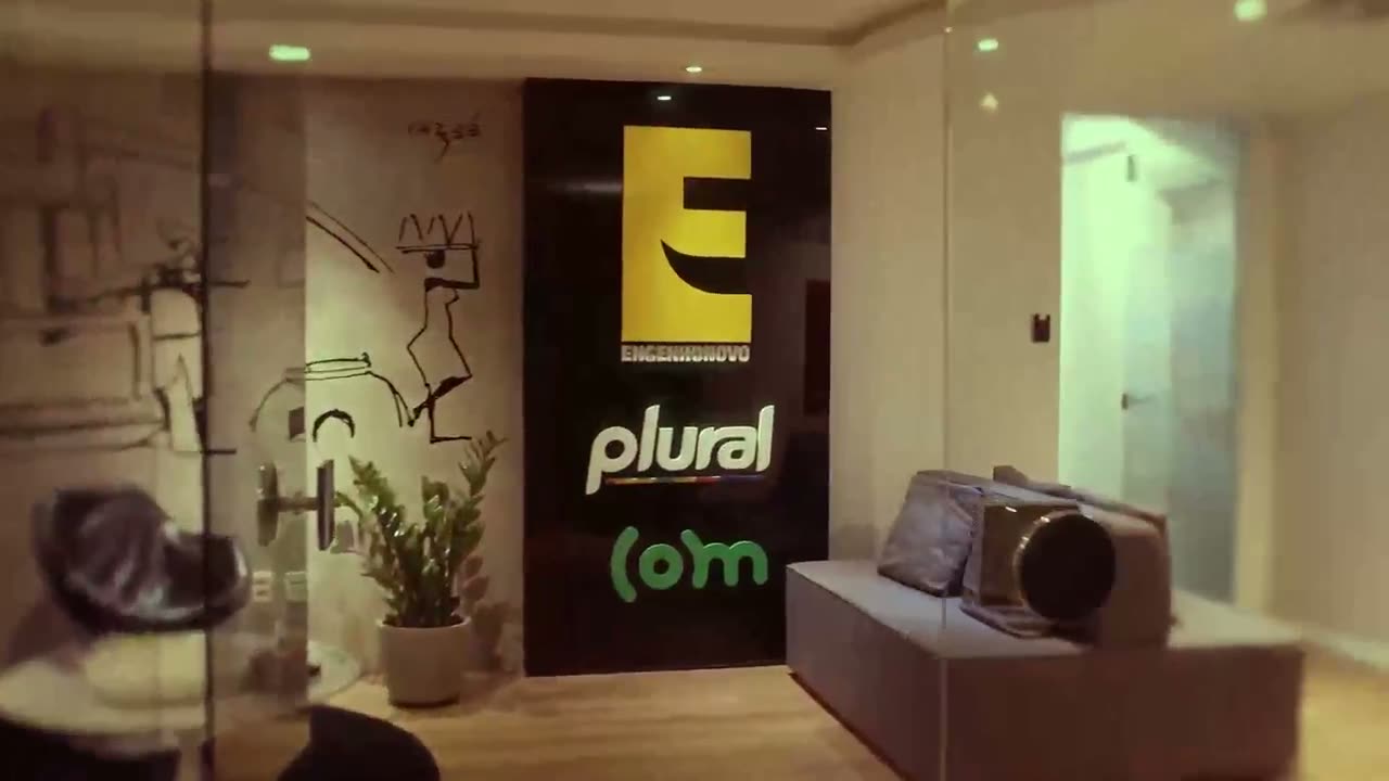 Sede do Grupo Engenho, com as marcas Engenho Novo, Plural e COM na parede e uma câmera sobre o sofá